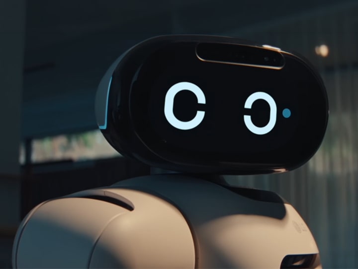 Close-up of a white humanoid robot with a black oval-shaped head display. The screen shows two large white circular eyes, and a small blue dot appears next to the right eye, suggesting a blinking or winking motion. The background is a softly blurred indoor space with warm lighting. 