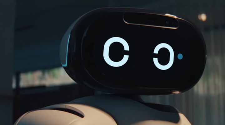 Close-up of a white humanoid robot with a black oval-shaped head display. The screen shows two large white circular eyes, and a small blue dot appears next to the right eye, suggesting a blinking or winking motion. The background is a softly blurred indoor space with warm lighting. 
