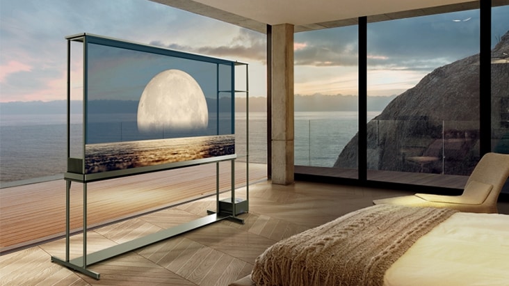 A transparent TV display stands in a luxurious bedroom with floor-to-ceiling windows overlooking the ocean and rocky cliffs at sunset. The see-through screen shows an image of a large moon rising over a calm sea. A bed with beige bedding is visible on the right side, and warm wooden flooring extends throughout the room.