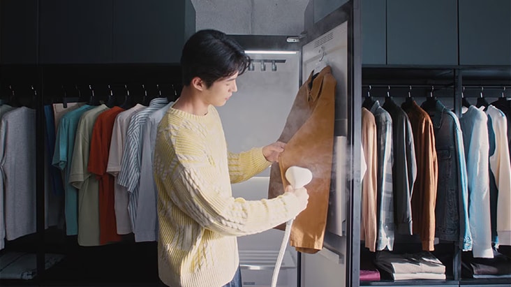 A young man wearing a yellow knit sweater stands in a walk-in closet, holding a brown leather jacket in front of an open clothing care appliance that emits steam. Rows of neatly hung shirts in various pastel colors line both sides of the closet.