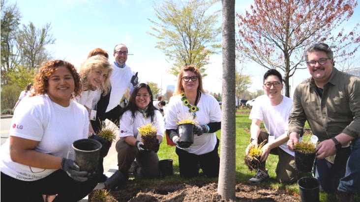 A group of smiling volunteers participating in a tree-planting and gardening activity on a sunny day. They are wearing white t-shirts and gloves, holding small potted plants and yellow grasses, posing together around a newly planted tree in an outdoor park or community area.