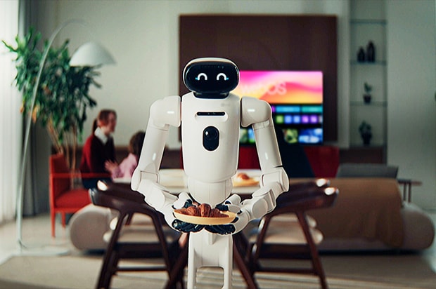A white humanoid robot stands in a warmly lit living room, holding a wooden plate with croissants in its hands. The robot has a rounded black face display showing two smiling curved eyes, giving it a friendly expression. In the blurred background, a woman and a child are seated at a dining table, and a large TV screen displays a colorful interface with the text "webOS" partially visible. A potted plant and shelves with decorative items are also visible in the background.