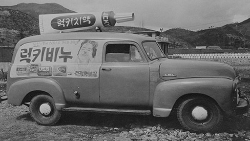 A vintage black and white photograph of an old delivery truck parked on a gravel road with mountains in the background. The side of the truck displays large Korean text reading "Lucky Soap" along with an illustration of a smiling woman's face and smaller product advertisements. On top of the truck is a large tube-shaped sign with Korean text reading "Lucky Toothpaste." The letters "GMC" are visible on the front of the truck.