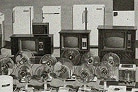 A vintage black and white photograph showing a display of home appliances arranged in rows. In the back row are several white refrigerators of various sizes. The middle row features wooden cabinet-style television sets with round CRT screens. The front row displays multiple electric fans with circular cage guards. An air conditioner unit is visible on the right side.