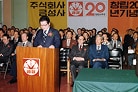 A photograph of a formal corporate ceremony with a man in a suit speaking at a wooden podium that displays the Geumseong (Gold Star) crown logo with Korean text reading "Geumseong." Behind him, rows of seated men in suits fill the audience. Banners on the wall show Korean text reading "Geumseong Corporation" and "20th Anniversary Celebration" along with a red circular emblem displaying "20" and Korean text reading "20th Anniversary."