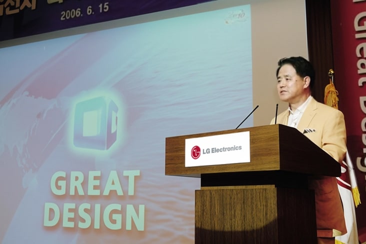 A photograph of a man in a beige jacket speaking at a wooden podium with the LG Electronics logo on the front. To his left, a large screen displays bold text reading "GREAT DESIGN" alongside an illustration of a square design icon and a wave-like graphic. The date "2006. 6. 15" is visible at the top of the screen. A South Korean flag is partially visible behind the podium.