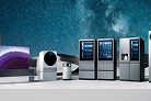 A product showcase featuring a lineup of premium LG home appliances arranged on a light platform against a deep blue cosmic background with stars and a galaxy. From left to right: a curved OLED TV displaying a purple coral-like image, a small white smart speaker, a sleek white robotic device, a black cylindrical speaker, two tall wine cellars with glass doors showing bottles inside, and a large stainless steel refrigerator with a transparent panel.
