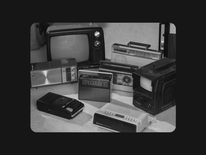 A vintage black and white photograph showing several hands working on assembling or inspecting a dark rectangular object, possibly an early electronic device or appliance component. The image has a grainy, archival film quality with rounded corners, framed against a black background. Multiple hands are visible from different angles, suggesting a collaborative workshop or factory setting.