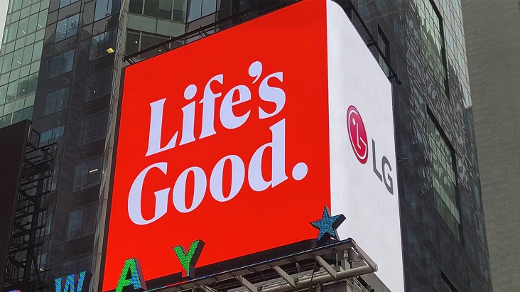 A photograph of a large outdoor digital billboard mounted on a modern glass-faced building in an urban setting, likely Times Square. The billboard displays a bold red background with large white text reading "Life's Good." along with the red and white LG logo on the right side. Other illuminated signs are partially visible at the bottom of the image.