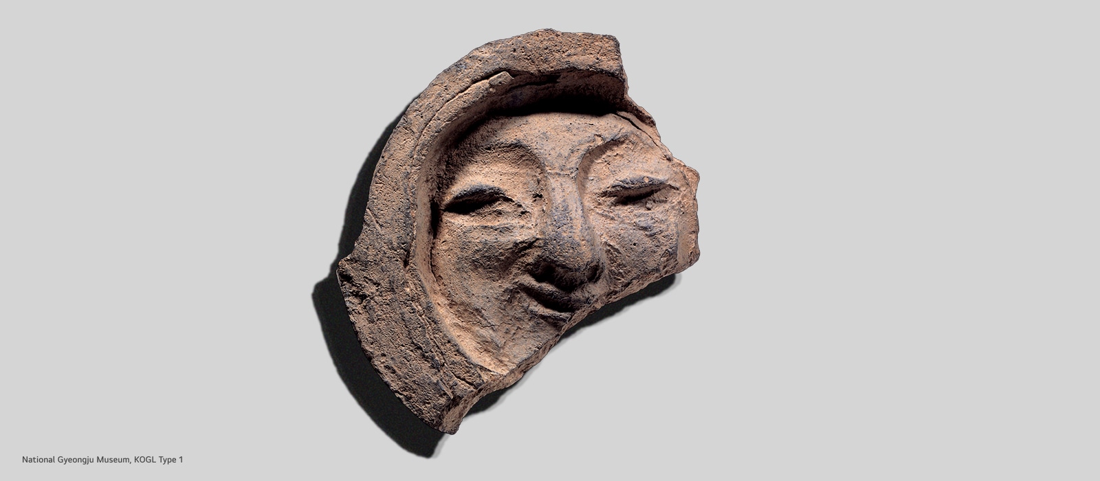 A close-up photograph of an ancient terracotta artifact depicting a smiling human face with closed eyes and curved mouth, set against a light gray background. The artifact appears weathered and broken at the edges, showing its archaeological age. Small text in the bottom left corner reads "National Gyeongju Museum, KOGL Type 1."