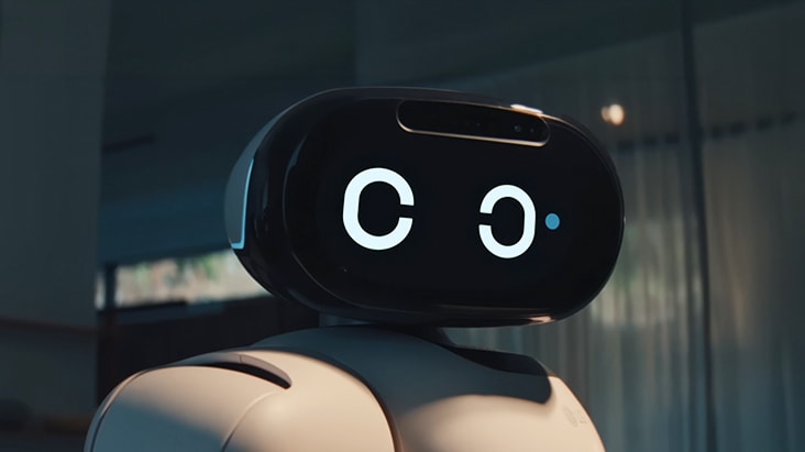 Close-up of a white humanoid robot with a black oval-shaped head display. The screen shows two large white circular eyes, and a small blue dot appears next to the right eye, suggesting a blinking or winking motion. The background is a softly blurred indoor space with warm lighting. 