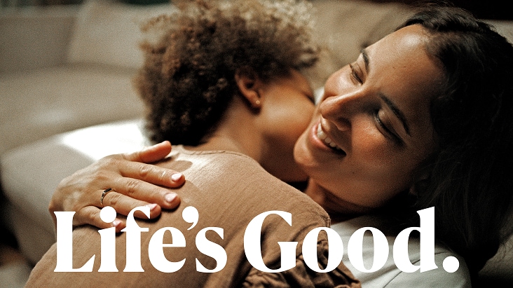 A smiling woman embracing a child indoors, with the text "Life's Good." overlaid at the bottom.