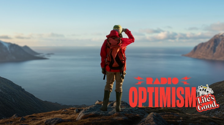 A promotional graphic with a scenic background of mountains and ocean. Bold red text reads "RADIO OPTIMISM" with lightning bolt icons on either side of "RADIO." Below the text is an illustration of a small white robot character holding a microphone, next to a red speech bubble containing the white text "Life's Good."