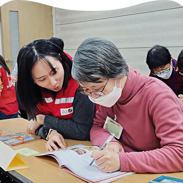 An LG School program scene where an LG staff member supports a senior participant at a welfare center, using an easy-to-read book to guide learning about home appliance usage
