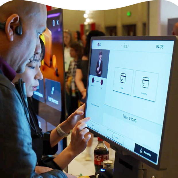 Two people use an LG kiosk with a sign language guide, selecting payment options and viewing a total of $10.00 on the touchscreen at an exhibition.
