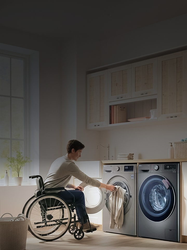 A person using a wheelchair loads laundry into a front-loading washing machine in a laundry room, with a dryer positioned beside it.