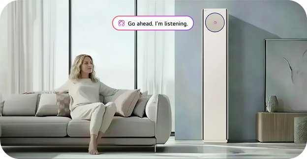 A woman sitting comfortably on a living room sofa, looking at a slim LG floor-standing air conditioner. A speech bubble with the text 'Go ahead. I'm listening.' and a voice recognition icon is displayed above the unit, indicating its smart voice control feature.