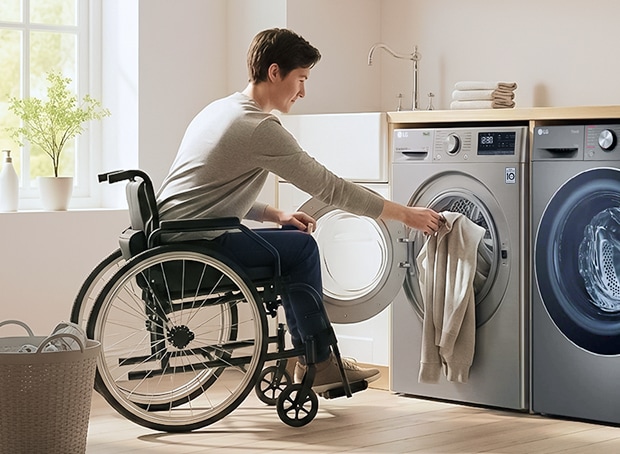 A person using a wheelchair loads laundry into a front-loading washing machine in a laundry room, with a dryer positioned beside it.