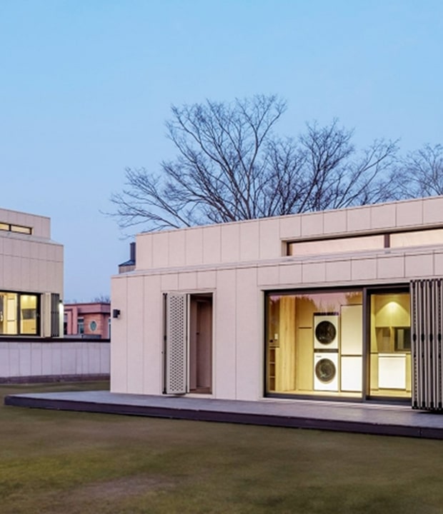 The exterior of the LG Smart Cottage, a modern smart home set on a lawn, with large glass windows revealing an interior space equipped with a washer and dryer.