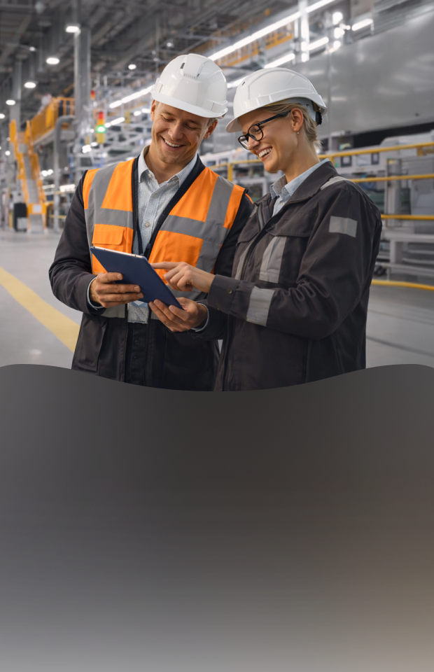 A spacious, brightly lit manufacturing plant interior equipped with industrial machinery. On the right, two workers wearing white hard hats, safety glasses, and high-visibility workwear are looking together at a tablet device.