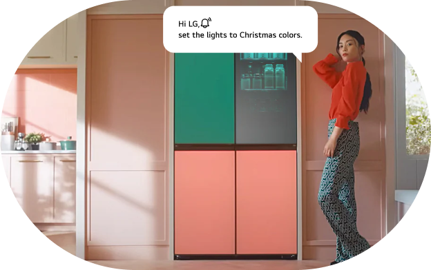 An image of the LG refrigerator AI voice command feature. A woman stands beside an LG refrigerator with a green upper section and pink lower section, with a voice command speech bubble displayed. Command: 'Hi LG! Set it to Christmas colors.'