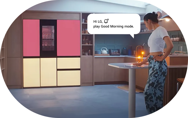 An image of the LG refrigerator AI voice command feature. A woman stands in a kitchen space with an LG refrigerator featuring a pink upper section and beige lower section, with a voice command speech bubble displayed. Command: 'Hi LG! Play Good Morning.'