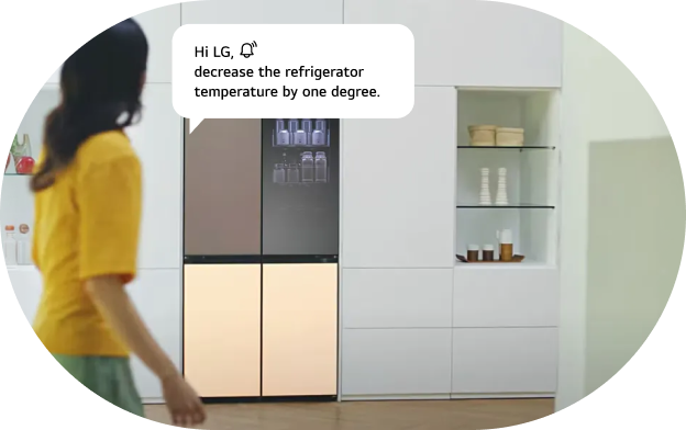 An image of the LG refrigerator AI voice command feature. A woman walks toward an LG refrigerator with a brown upper section and beige lower section, with a voice command speech bubble displayed. Command: 'Hi LG! Lower the refrigerator temperature by 1 degree.'