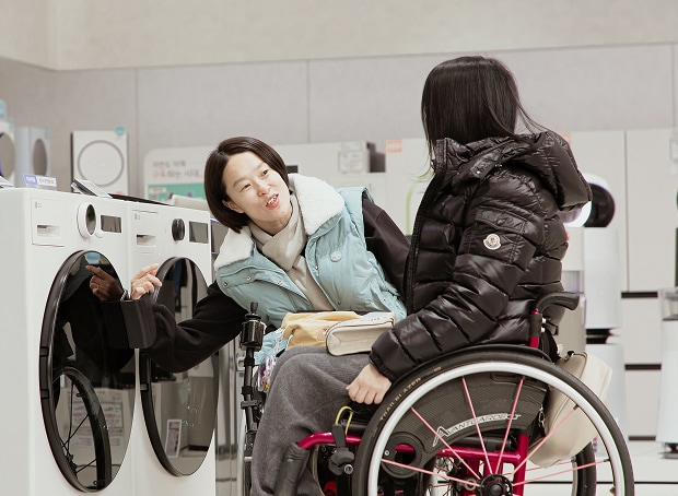 A person using a wheelchair loads laundry into a front-loading washing machine in a laundry room, with a dryer positioned beside it.
