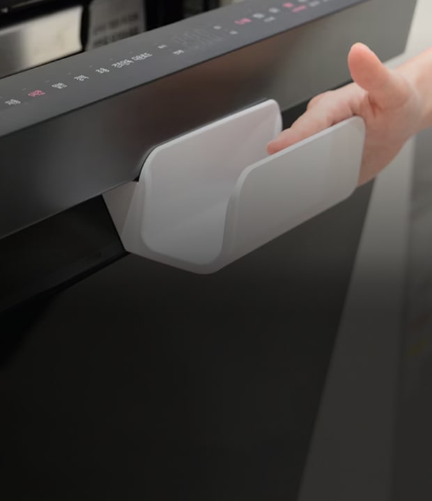 A close-up photo of a dark-colored LG dishwasher built into the lower section of a white kitchen counter, with its door slightly open. A person's hand reaches in from the right, gripping the white U-shaped auxiliary handle mounted on the top of the dishwasher door as if about to move it. On the left side of the black control panel along the top of the door, the LG logo is clearly visible, with a row of indicator lights and function buttons arranged beside it. Product information stickers are attached to the interior of the dishwasher and the lower front of the door, overall showing a clean, modern kitchen appliance in use.