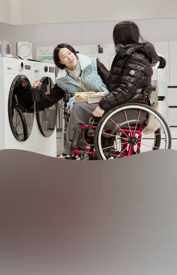 A person using a wheelchair loads laundry into a front-loading washing machine in a laundry room, with a dryer positioned beside it.