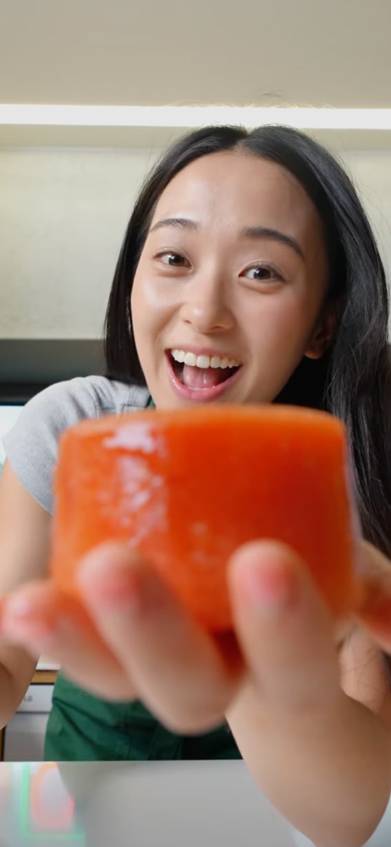 Woman with long dark hair smiling and holding a piece of sushi close to the camera.