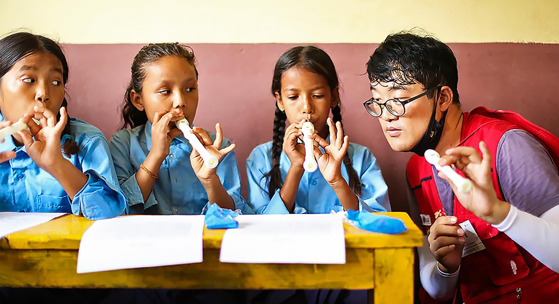 A LG volunteer wearing a red vest is teaching three children how to play the recorder.