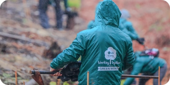 A LG employee wearing a green jacket with Green world written on it is seen from behind, holding a shovel on a dirt road.