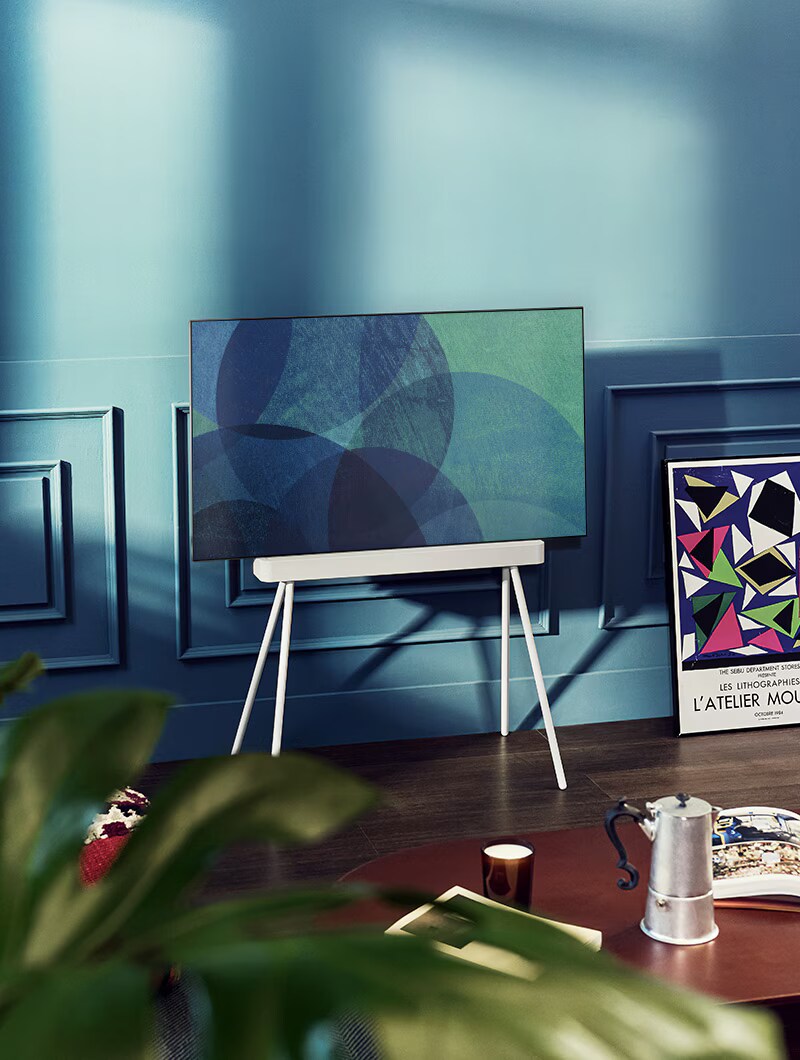 Telewizor OLED LG na podstawie Gallery Stand znajduje się w nowoczesnym szarym pokoju naprzeciwko okna wychodzącego na ośnieżony las. Telewizor OLED LG na podstawie Floor Stand znajduje się w kolorowym pokoju dziennym z pastelowymi i drewnianymi meblami.