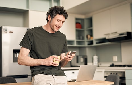 En kvinna sitter på en grå soffa i ett ljust och soligt vardagsrum medan hon tittar på sin mobiltelefon. Stora gröna krukväxter omger rummet, och ett runt soffbord med en bricka, en liten kaktus i kruka och en mugg står i förgrunden. Mjukt naturligt ljus strömmar in genom höga fönster med vita gardiner och skapar en lugn och mysig atmosfär.
