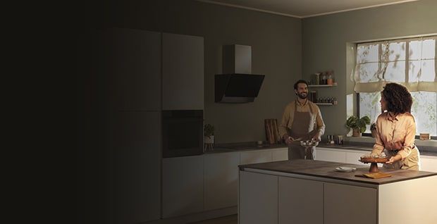 Modernt kök med en man och en kvinna som ler och pratar vid en köksö. Köket har stilrena LG-vitvaror, mjuk belysning och stora fönster som släpper in naturligt ljus. LG- och ThinQ-logotyper syns till vänster.