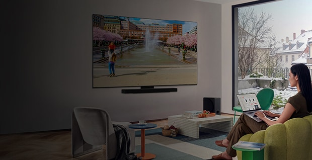 LG platt smart-TV monterad på väggen i ett vardagsrum, som visar en färgstark stadsscen, medan en person sitter i en grön soffa och tittar.