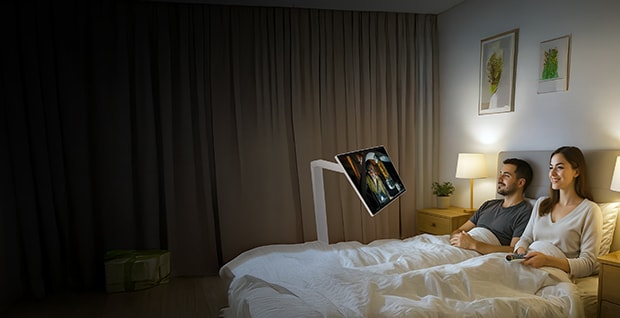 Ett par ligger i sängen och tittar på en LG Smart Monitor som är monterad på en Swing-arm framför dem. Rummet är mjukt upplyst med en sänglampa, och skärmen visar färgglatt innehåll. Båda ser avslappnade ut. Längst ner i bilden syns logotyperna för LG och Swing Smart Monitor.