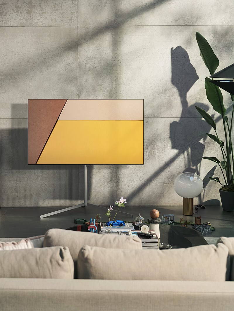 Tivi LG OLED trong một không gian sống màu xám hiện đại trên cửa sổ hiển thị một hồ bơi. Tivi LG OLED trong một không gian sống trung tính với gạch phơi sáng, các chi tiết bằng gỗ và cây xanh.