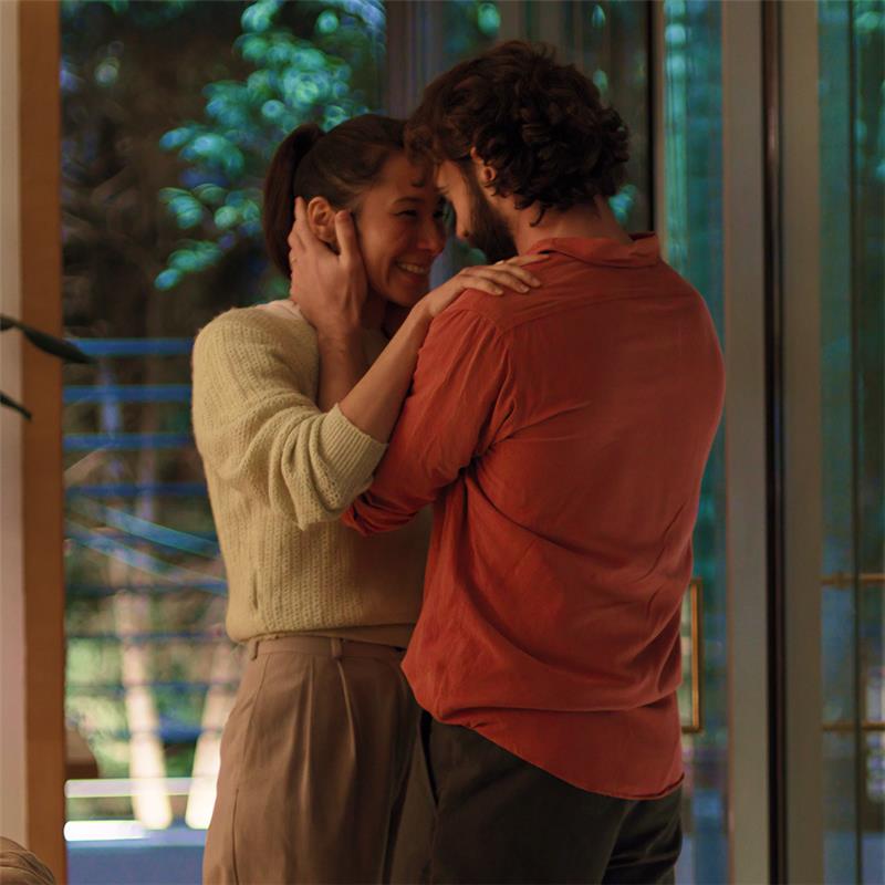 A man and woman share a warm embrace in a cozy living room, surrounded by soft lighting and comfortable furnishings.