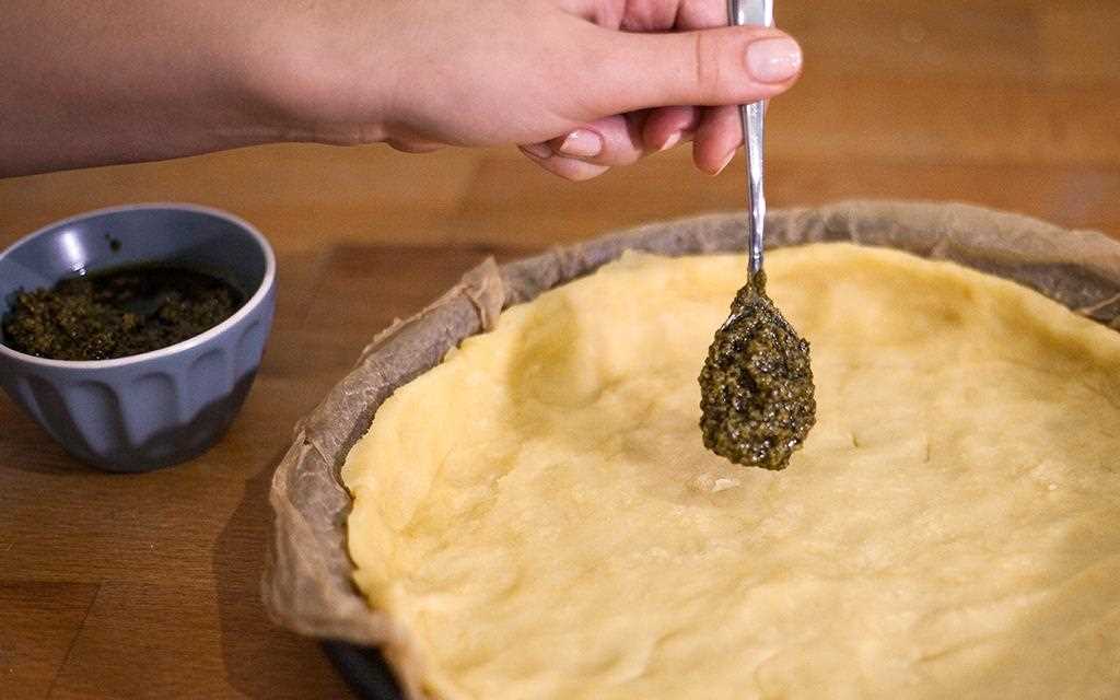 Oliven-Pesto wird auf dem Teig eines Tomaten-Quiches verteilt