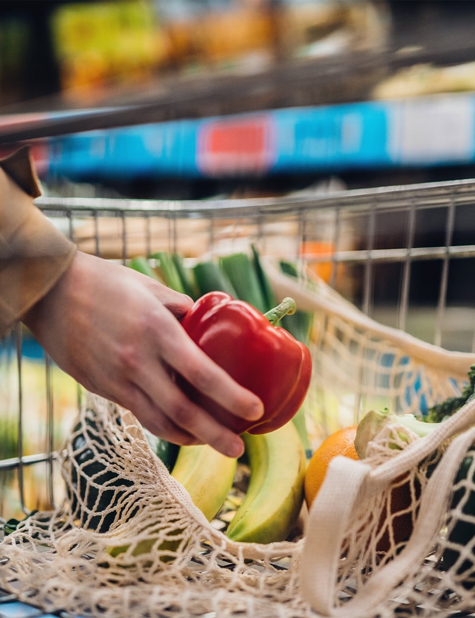 A grocery store shopper's hand is putting a red bell pepper into a reusable produce bag