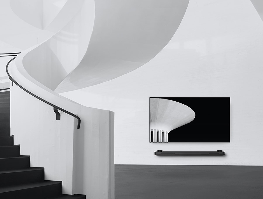 Ein Schwarz-Weiß-Bild von dem lg signature OLED TV, das links der Wand hängt und links davon befindet sich eine Wendeltreppe