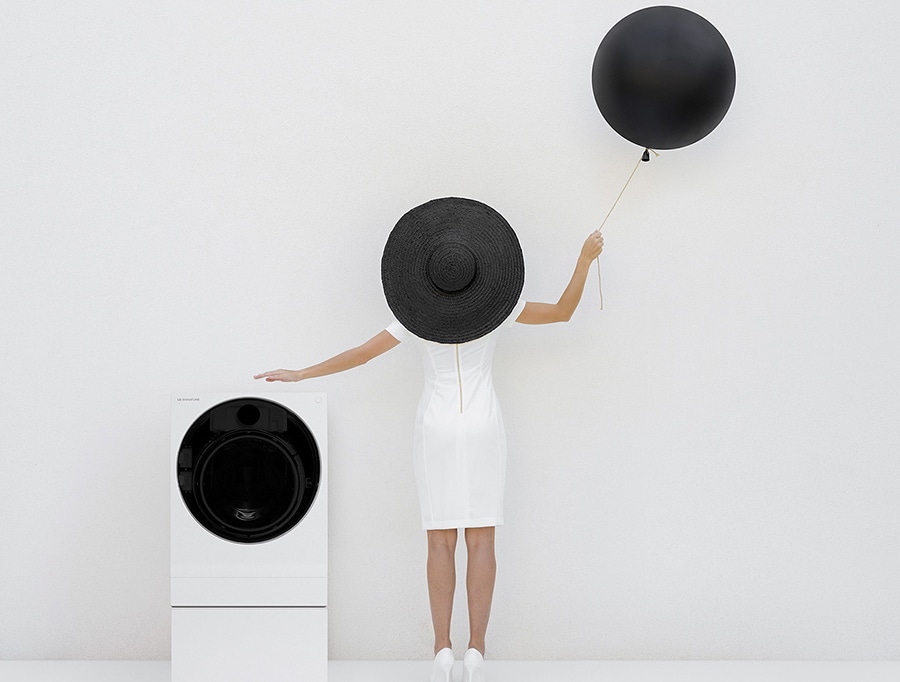 Die Waschmaschine von lg signature befindet sich unten links in dem gesamten weißen Bereich, und rechts in der Mitte steht Anna mit einem weißen Einteiler und trägt einen schwarzen Hut und Ballon in der rechten Hand
