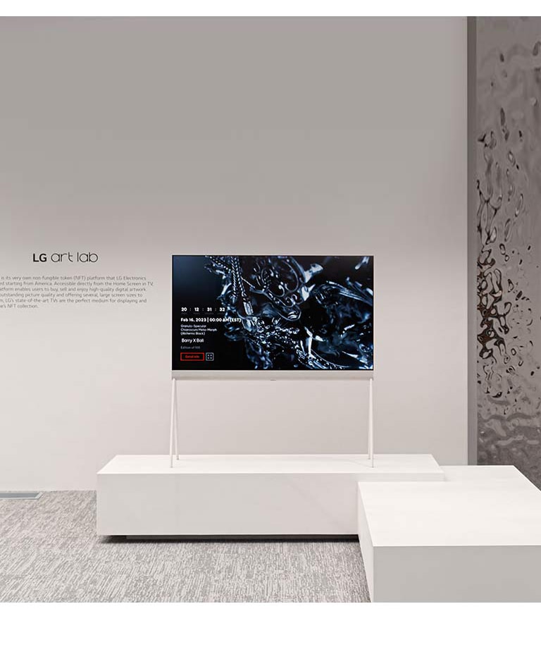 Et billede af et staffeli i et hvidt rum viser et digitalt kunstværk af en sort skulptur på skærmen. En sølvfarvet fysisk skulptur til højre for TV’et viser et spejlbillede af rummet.