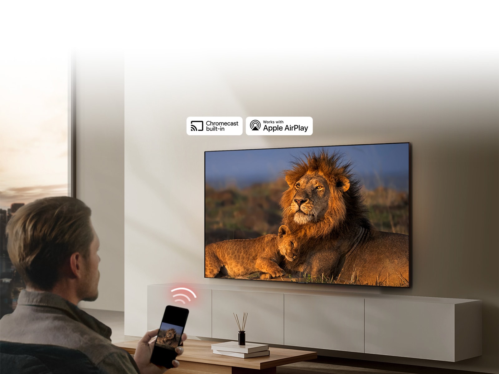 Un téléviseur LG fixé au mur d’un salon, montrant un lion et un lionceau. Un homme est assis au premier plan, un smartphone à la main affichant la même image de lions. Un graphique représentant trois barres incurvées rouge néon est affiché juste au-dessus du smartphone pointant vers le téléviseur.