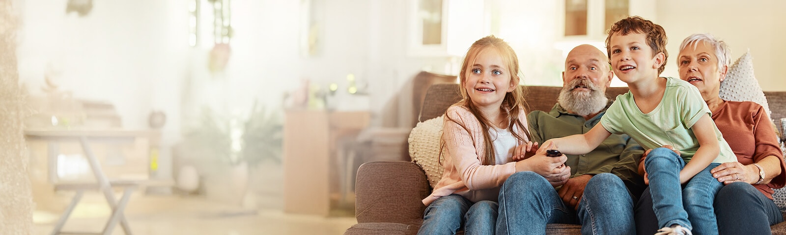 Une famille avec des enfants et leurs grands-parents sont assis ensemble sur un canapé dans un salon lumineux, tenant une télécommande tout en regardant la télévision.