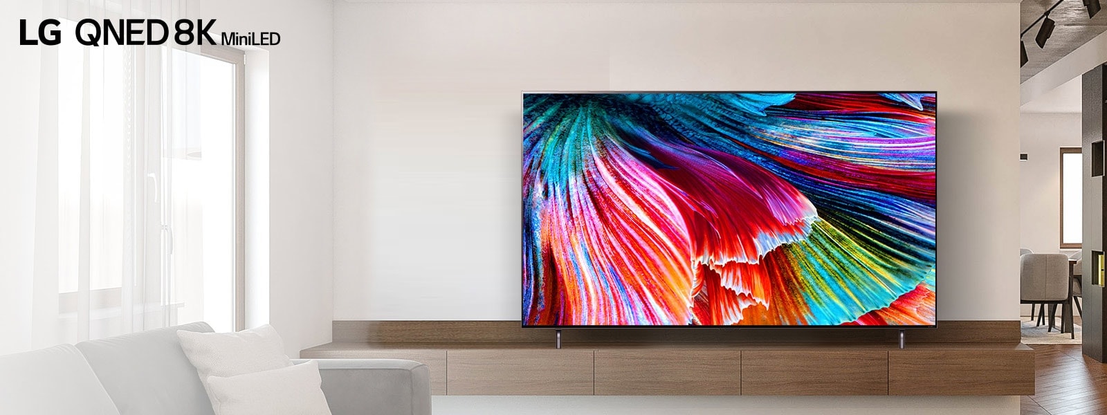 Téléviseur QNED MiniLED installé sur une armoire en bois contre un mur lumineux dans un intérieur moderne et lumineux. L’écran affiche une image en gros plan de pétales multicolores qui se déploient.