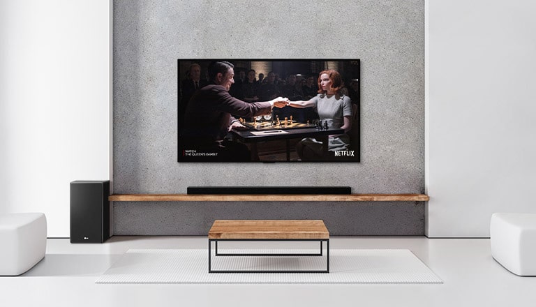 A subwoofer, and a soundbar, and TV are in a white living room. A woman and a man are playing chess on TV screen.