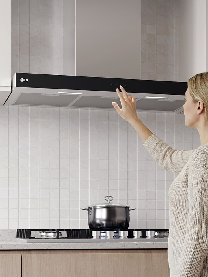 A woman adjusting the touch controls of a built-in hood above a cooktop in a modern kitchen with a white tile wall.
